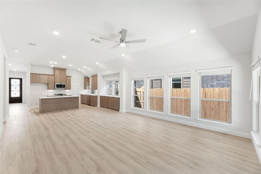 Open living area with light wood floors, vaulted ceiling, ceiling fan, and kitchen with brown cabinets and island in background Open living area with light wood floors, vaulted ceiling, ceiling fan, and kitchen with brown cabinets and island in background