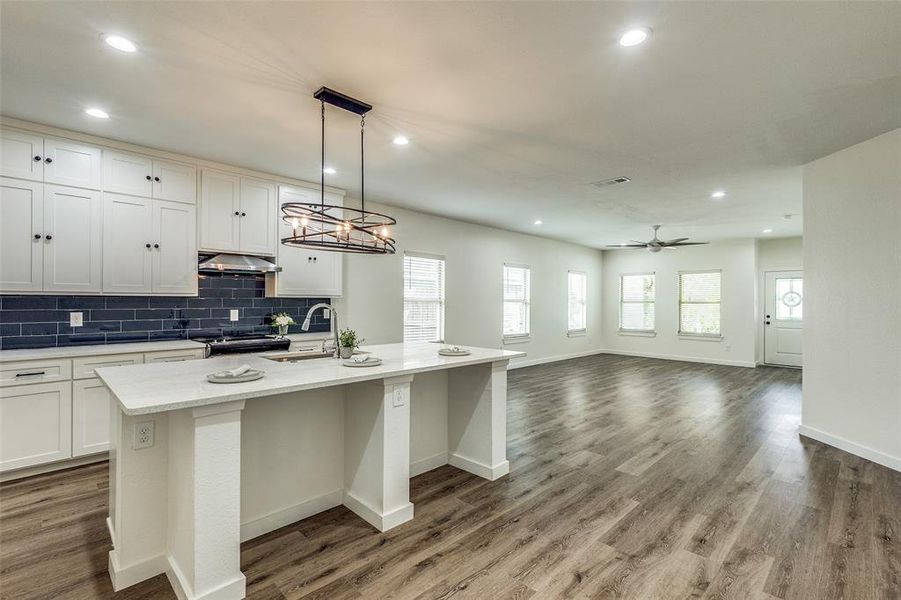 Kitchen with decorative backsplash, a kitchen island with sink, ceiling fan with notable chandelier, wood-type flooring, and decorative light fixtures