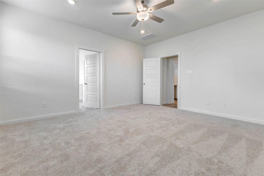 Another angle of the primary suite highlights the generous room size, ceiling fan, soft carpet, recessed lighting, and direct access to both the en-suite bath and main living areas. The neutral finishes create a fresh blank canvas for a comfortable bedroom layout and relaxing daily routine.