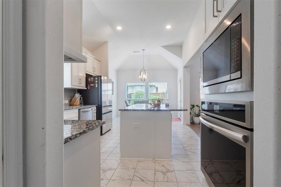 Kitchen with stainless steel appliances, dark stone countertops, pendant lighting, light marble finish flooring, and a center island