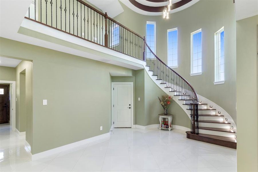 This elegant home entryway features a grand curved staircase with dark wood steps and wrought iron railings. The high ceiling and tall windows provide ample natural light, while the light green walls and glossy white floors add a touch of modern sophistication.