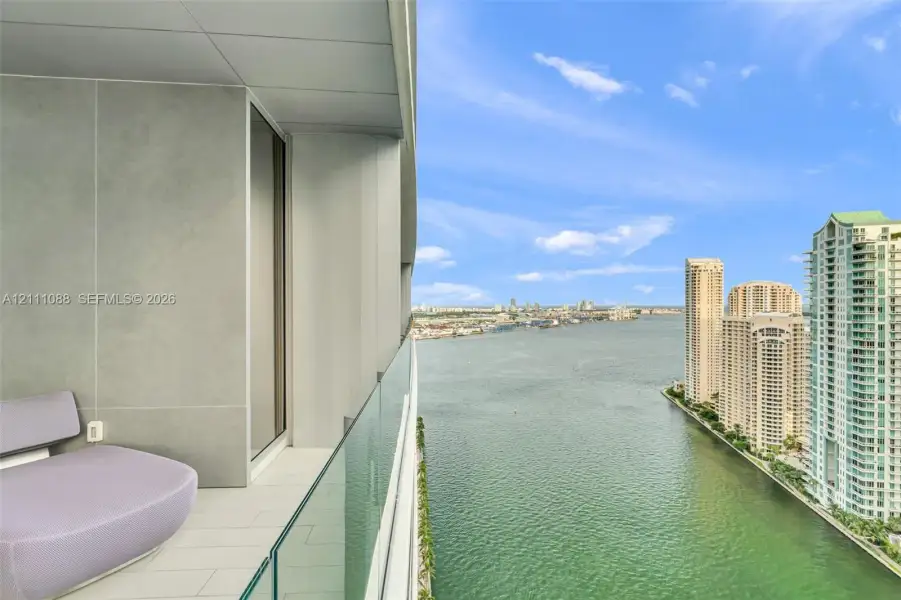 Private balcony featuring large-format tile flooring, concrete-finish wall panels, and a frameless glass railing overlooking the waterway Private balcony featuring large-format tile flooring, concrete-finish wall panels, and a frameless glass railing overlooking the waterway