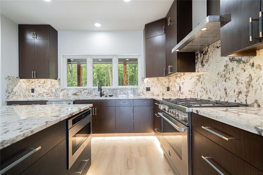 Kitchen with stainless steel appliances, wall chimney range hood, light stone counters, backsplash, and dark brown cabinets Kitchen with stainless steel appliances, wall chimney range hood, light stone counters, backsplash, and dark brown cabinets