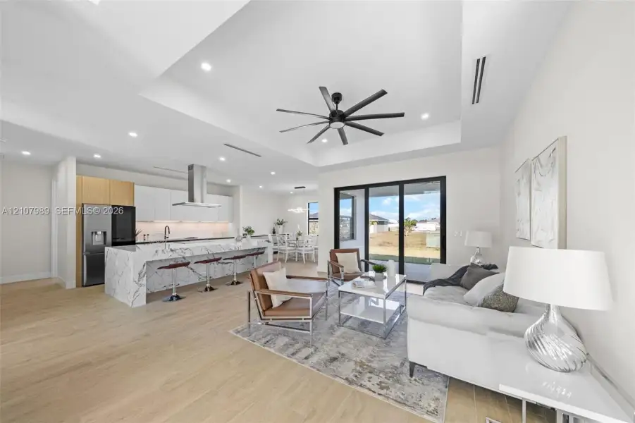 Tray ceiling with multi-blade ceiling fan, expansive kitchen island with stone-finish waterfall countertop, wood-finish flooring, and multi-panel sliding glass doors Tray ceiling with multi-blade ceiling fan, expansive kitchen island with stone-finish waterfall countertop, wood-finish flooring, and multi-panel sliding glass doors