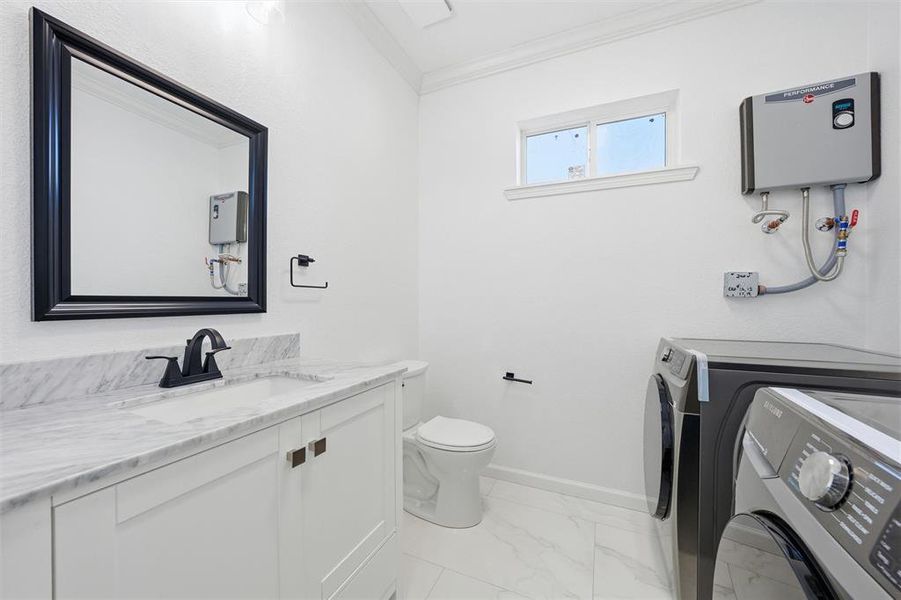Half bath featuring light marble finish floors, ornamental molding, water heater, vanity, and washing machine and dryer Half bath featuring light marble finish floors, ornamental molding, water heater, vanity, and washing machine and dryer
