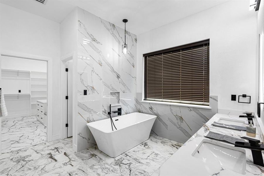 Bathroom featuring a walk in closet, a freestanding tub, double vanity, light marble finish flooring, and stone wall