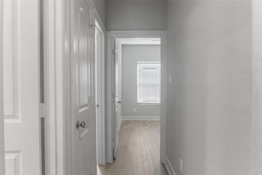 A bright, narrow hallway with light gray walls and wood flooring leads to a room with a window fitted with blinds. There are multiple doors along the passage.