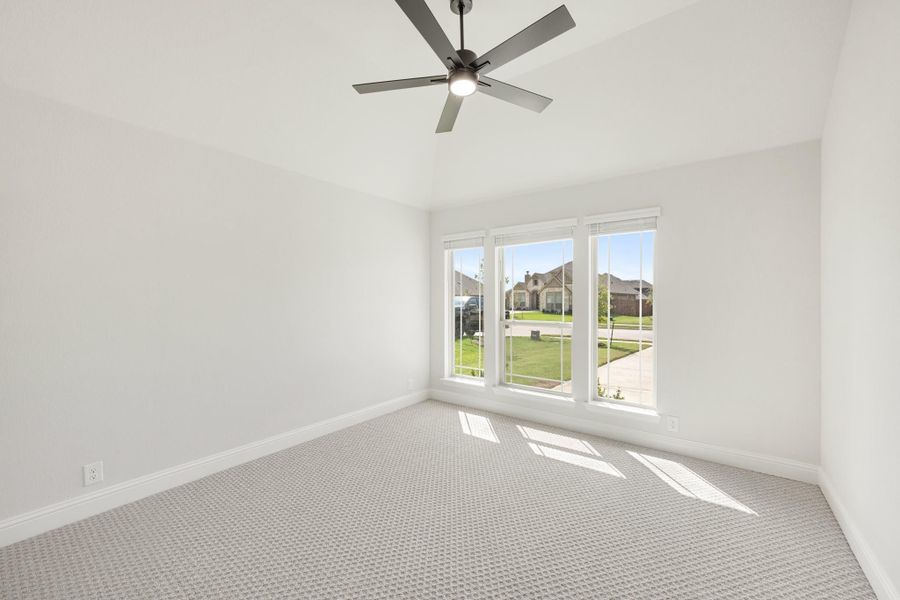 Empty bedroom with light gray carpet, ceiling fan, and large windows overlooking a neighborhood street.