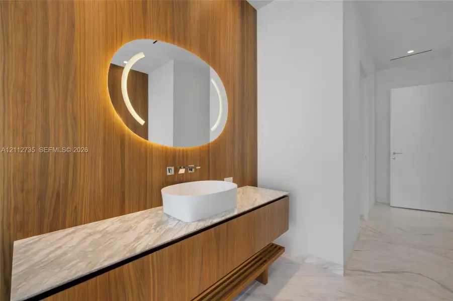 Floating wood-finish vanity with a stone countertop, white vessel sink, wall-mounted faucet, and oval backlit mirror set against a floor-to-ceiling wood-paneled wall