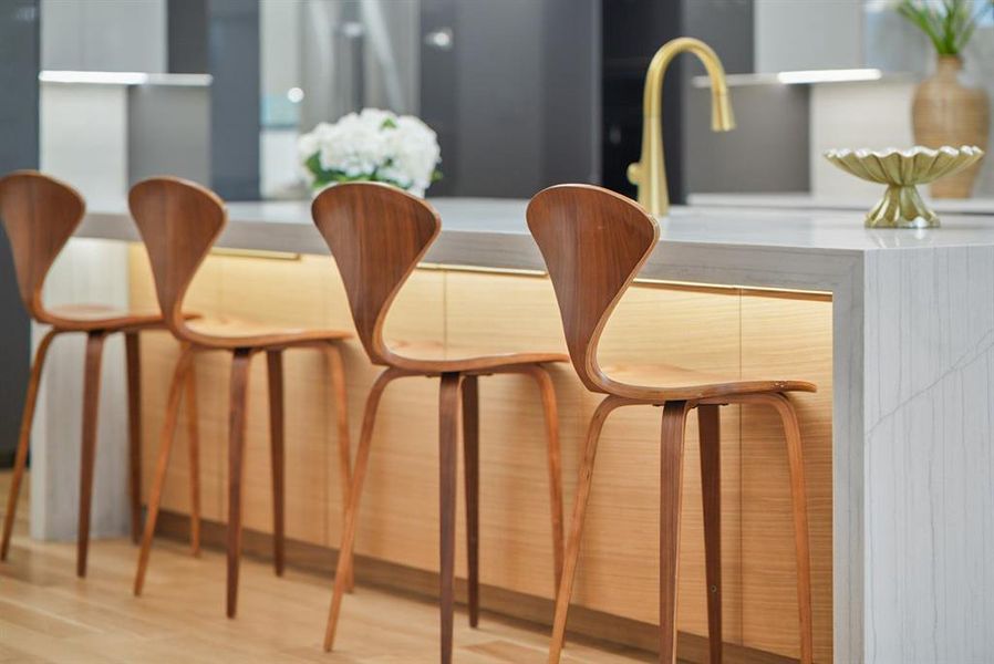 Kitchen island featuring a waterfall-edge countertop, under-cabinet lighting, light wood-finish cabinetry, and a gold-finish faucet