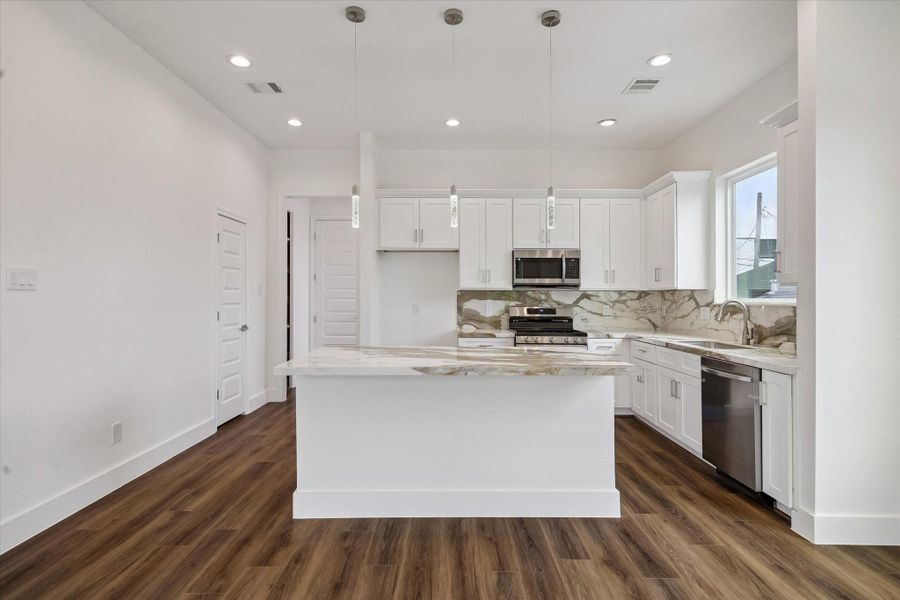 Designed for both function and style, the kitchen centers around an impressive island with abundant workspace and storage. White cabinetry extends across the rear wall, framed by dramatic stone-look counters and backsplash, while stainless steel appliances complete the modern look. High ceilings, recessed lighting, pendant fixtures, and rich wood-look flooring give the room an airy, upscale feel. The generous proportions create plenty of room for cooking, gathering, and casual dining. Designed for both function and style, the kitchen centers around an impressive island with abundant workspace and storage. White cabinetry extends across the rear wall, framed by dramatic stone-look counters and backsplash, while stainless steel appliances complete the modern look. High ceilings, recessed lighting, pendant fixtures, and rich wood-look flooring give the room an airy, upscale feel. The generous proportions create plenty of room for cooking, gathering, and casual dining.