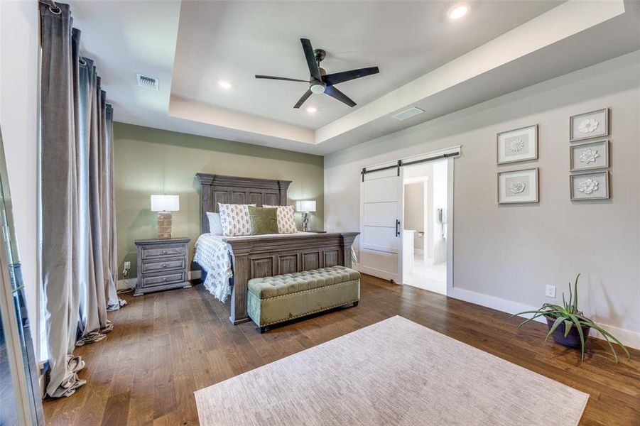 Bedroom featuring a barn door, a tray ceiling, dark wood-type flooring, a ceiling fan, and recessed lighting