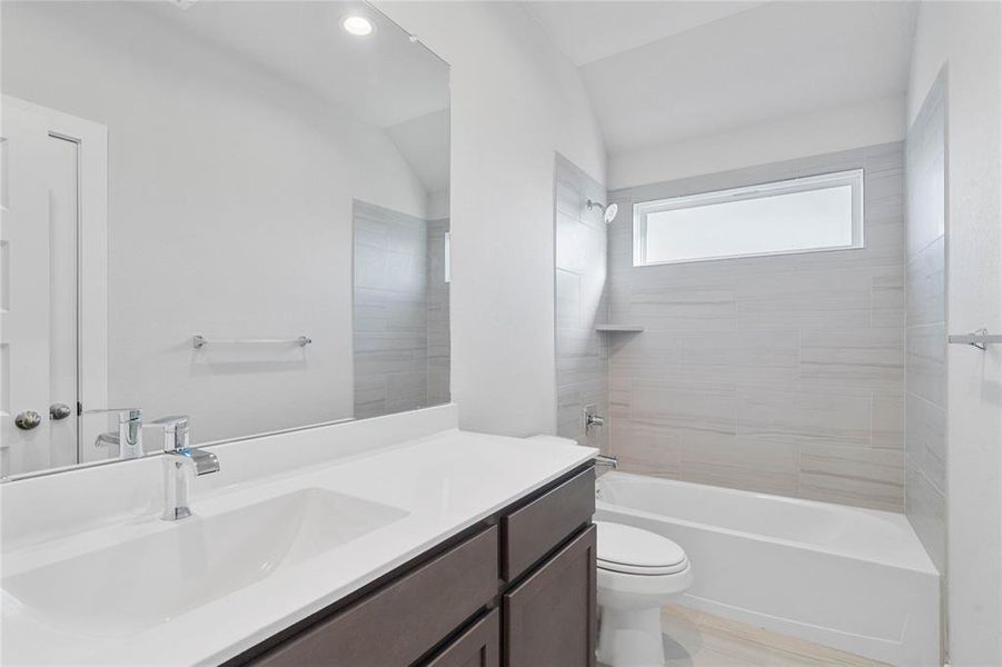 Secondary bath features tile flooring, bath/shower combo with tile surround, stained wood cabinets, beautiful light countertops, mirror, dark, sleek fixtures and modern finishes!