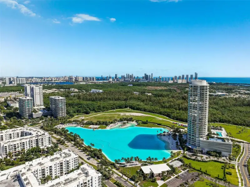 Aerial perspective of a high-rise residential tower featuring a large man-made lagoon with a sandy shoreline, surrounding parkland, and coastal city skyline