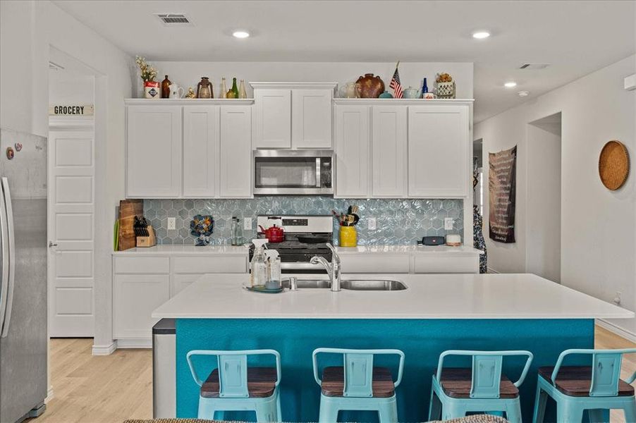 Kitchen featuring light wood-type flooring, backsplash, stainless steel appliances, a center island with sink, and white cabinetry