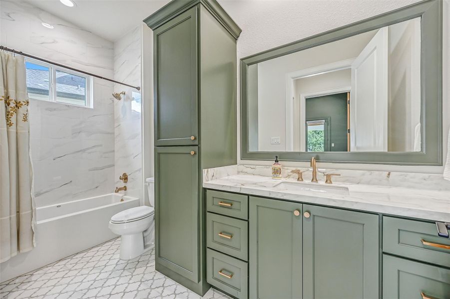 A nearby full bath features a custom sage-green vanity with abundant storage, coordinating framed mirror, stone-look countertop and a tub/shower surrounded by marble-look tile extending to the ceiling.