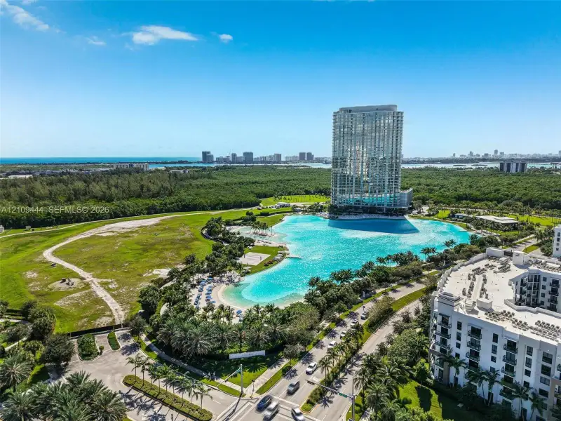 Aerial perspective featuring a large crystalline lagoon, expansive landscaped grounds, mature palm trees, and proximity to the coastline