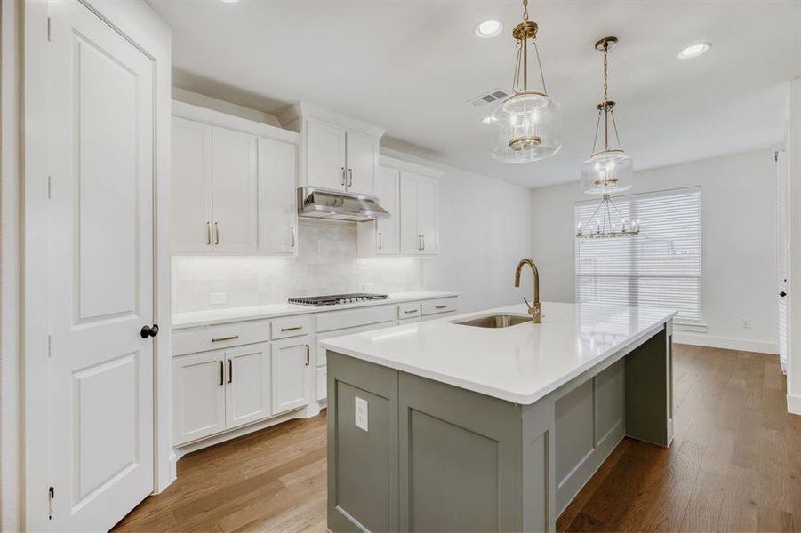 Modern kitchen featuring a large island with sink and bronze-finish faucet, white shaker cabinetry with under-cabinet lighting, stainless steel range hood, and wood-finish flooring