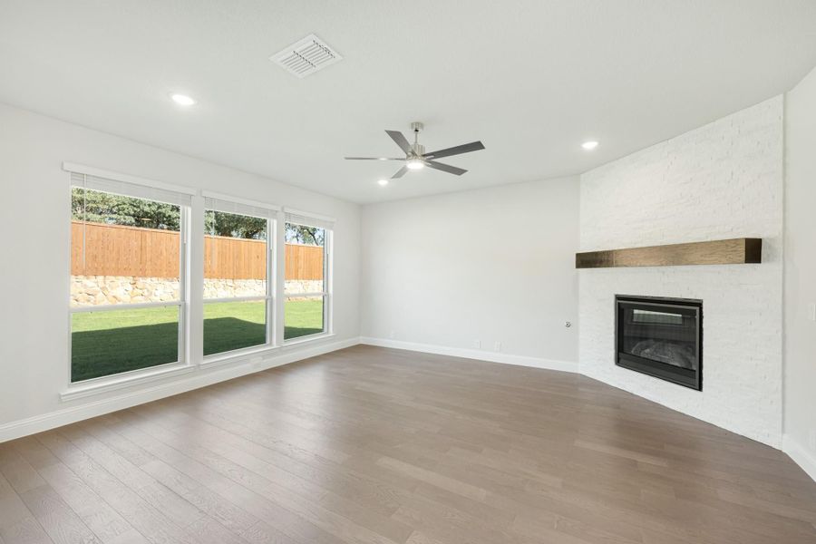 Open living room with hardwood floors, white brick fireplace, ceiling fan, and large windows overlooking backyard