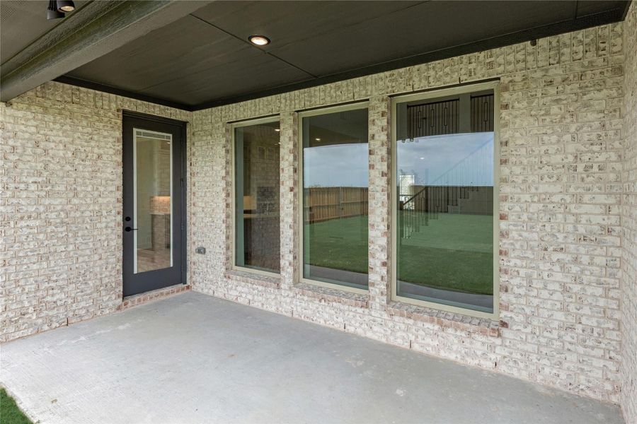 Covered back patio with concrete floor, light brick exterior, dark door, and two large windows overlooking backyard