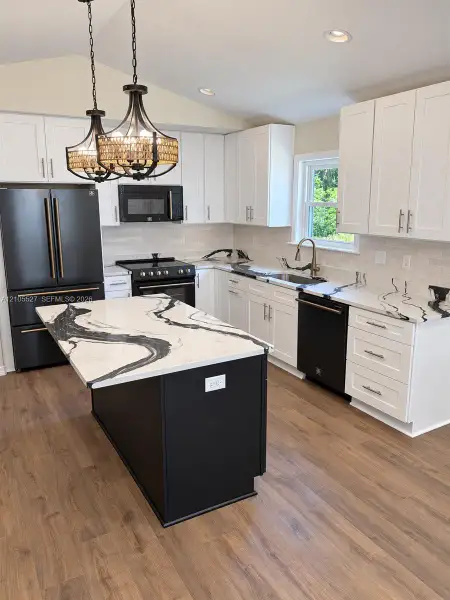 Kitchen featuring white shaker cabinetry, black appliances, a central island with a waterfall countertop, wood-finish flooring, and two decorative pendant lights Kitchen featuring white shaker cabinetry, black appliances, a central island with a waterfall countertop, wood-finish flooring, and two decorative pendant lights