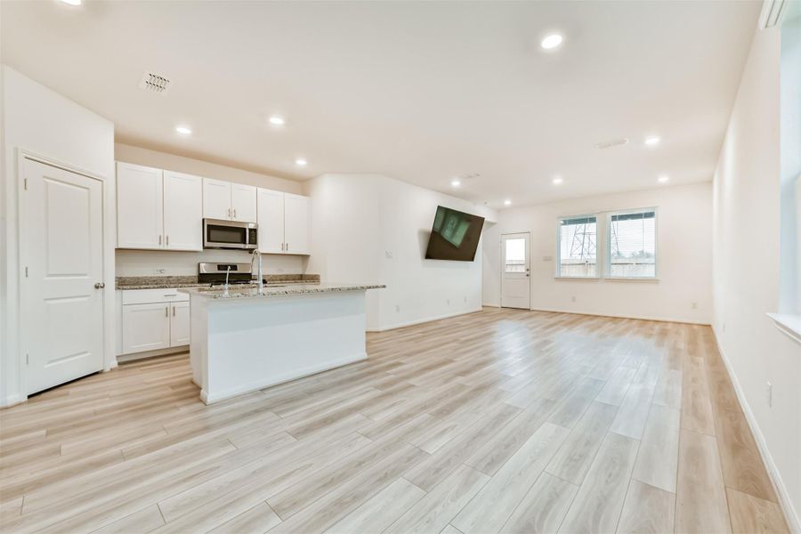Bright, open-concept living space featuring a modern kitchen with white cabinets, granite countertops, and a central island. The area is complemented by recessed lighting, large windows, and wood flooring, creating an inviting atmosphere.
