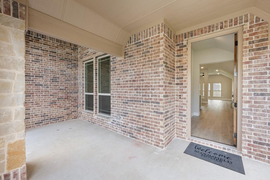 Covered front porch with brick exterior, open entry door, and welcome mat leading into hardwood-floored interior.