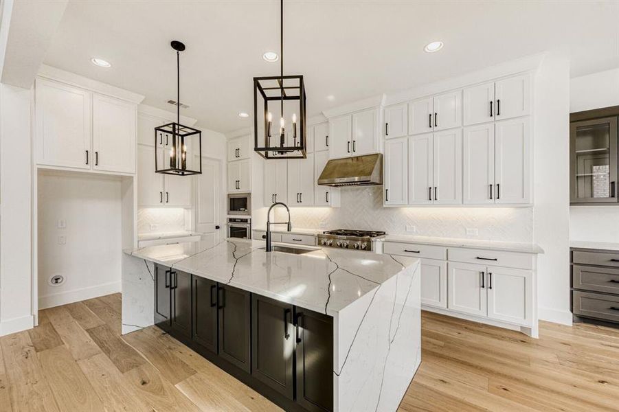 Two tone kitchen featuring two tone cabinets, light stone countertops, backsplash, a center island with sink, and light wood-style flooring