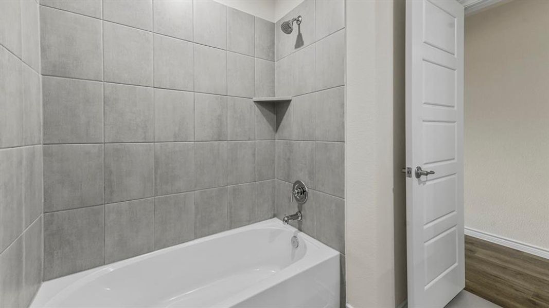 Tub and shower combination featuring large format grey wall tiles, a built-in corner shelf, and brushed nickel fixtures