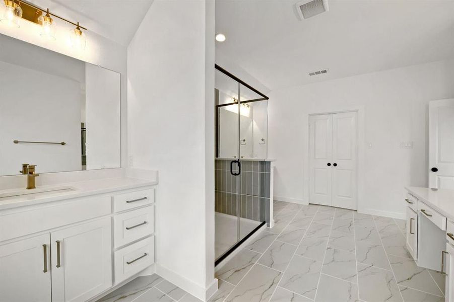 Spacious bathroom featuring a frameless glass shower enclosure, a dual vanity with white cabinetry and light countertops, modern gold-tone fixtures, and large format floor tiles Spacious bathroom featuring a frameless glass shower enclosure, a dual vanity with white cabinetry and light countertops, modern gold-tone fixtures, and large format floor tiles