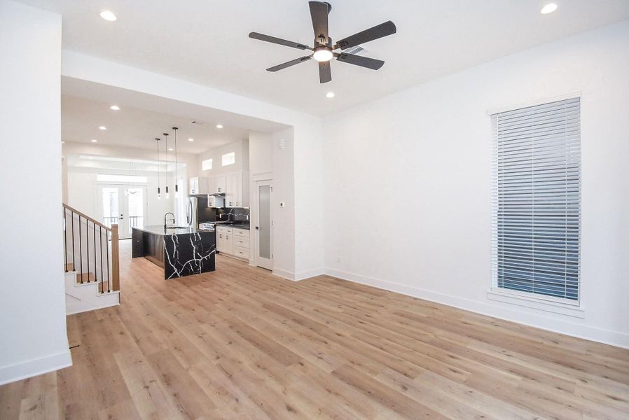 This photo showcases a spacious, open-concept living area with light wood flooring and a ceiling fan. The modern kitchen features a stylish black marble island, sleek white cabinets, and stainless steel appliances. Large windows and recessed lighting add brightness, while a staircase leads to the upper level.