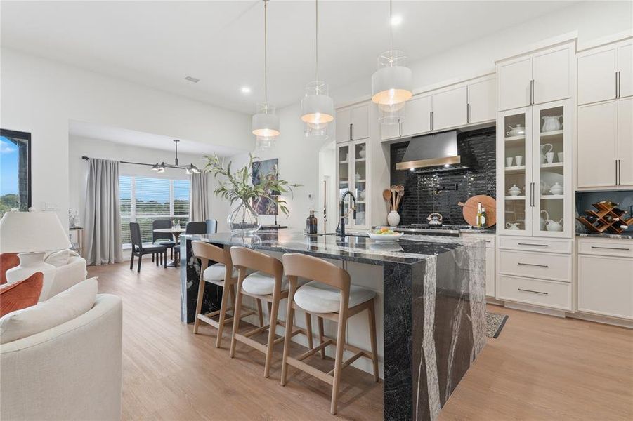 Gourmet kitchen featuring a large waterfall island with seating, dark tile backsplash, stainless steel range hood, ample cabinetry with glass-front accents, and light wood-finish flooring