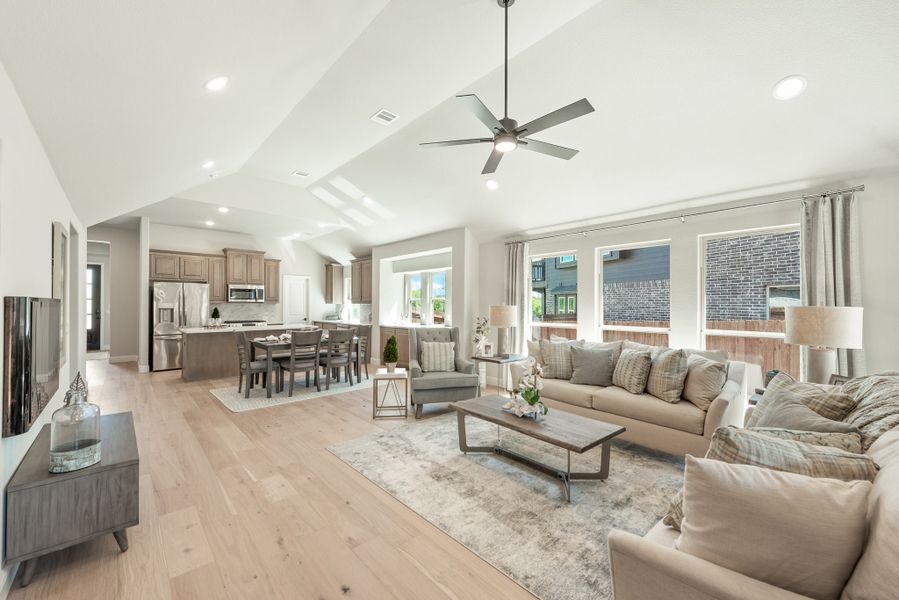 Open-concept living room with vaulted ceiling, ceiling fan, hardwood floors, and kitchen visible in background