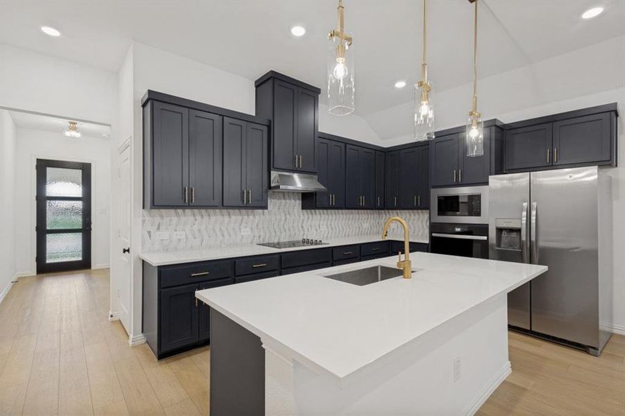 Contemporary kitchen featuring dark cabinetry, white countertops, stainless steel appliances, a geometric tile backsplash, and a central island with an undermount sink and gold-tone faucet