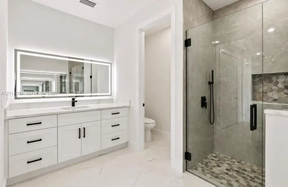 Bathroom featuring a white shaker-style vanity with solid-surface countertop, an integrated LED-backlit mirror, a walk-in shower with large-format tile surround, matte black hardware, and hexagonal tile flooring Bathroom featuring a white shaker-style vanity with solid-surface countertop, an integrated LED-backlit mirror, a walk-in shower with large-format tile surround, matte black hardware, and hexagonal tile flooring