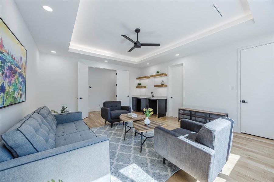 Living room with light wood-style flooring, a tray ceiling, a ceiling fan, recessed lighting, and bar with sink Living room with light wood-style flooring, a tray ceiling, a ceiling fan, recessed lighting, and bar with sink