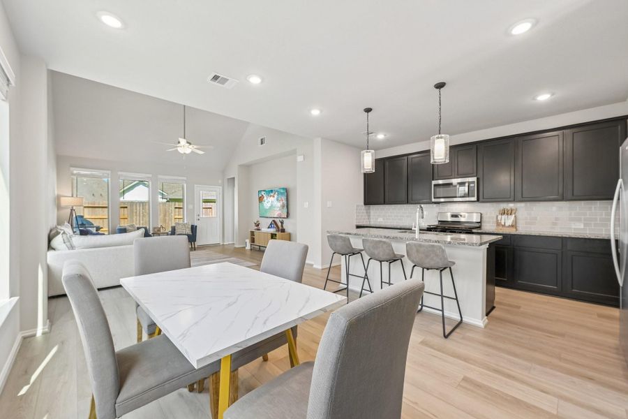 Bright and open living space with a modern kitchen featuring dark cabinetry, stainless steel appliances, and a spacious island with seating, seamlessly flowing into the airy dining and living areas.