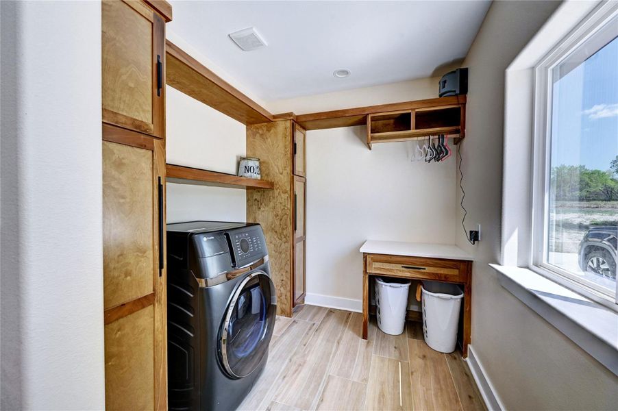 This is a modern laundry room with a sleek black washer, ample wood cabinetry, and a folding station. It features a large window allowing natural light, and has bins and hanging space for convenience.