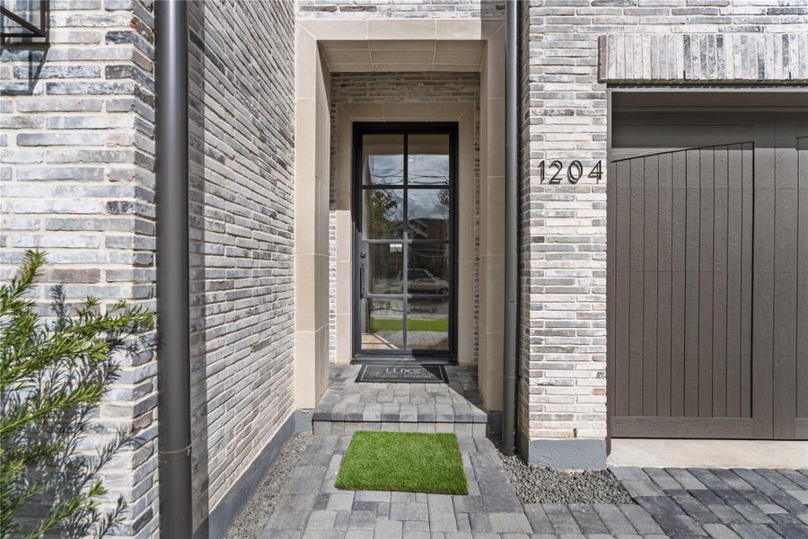 The entry is bolstered by a custom limestone surround, subtly contrasting the gray toned brick and framing the space. Pavers line the path and provide a rustic, Old World feel. The pivoting black steel door previews the splendor to come!