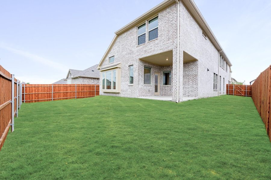 Two-story brick home rear exterior with covered patio, large grassy backyard, and wood privacy fence