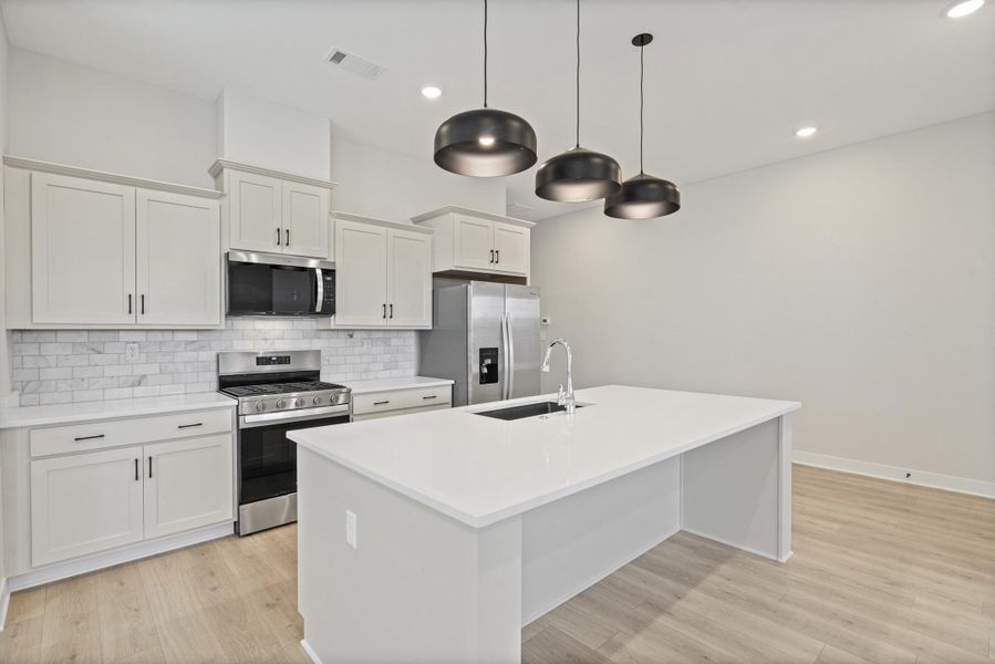 Modern kitchen featuring light-toned cabinetry with dark hardware, stainless steel appliances, a subway tile backsplash, a large center island with an undermount sink, and wood-finish flooring Modern kitchen featuring light-toned cabinetry with dark hardware, stainless steel appliances, a subway tile backsplash, a large center island with an undermount sink, and wood-finish flooring