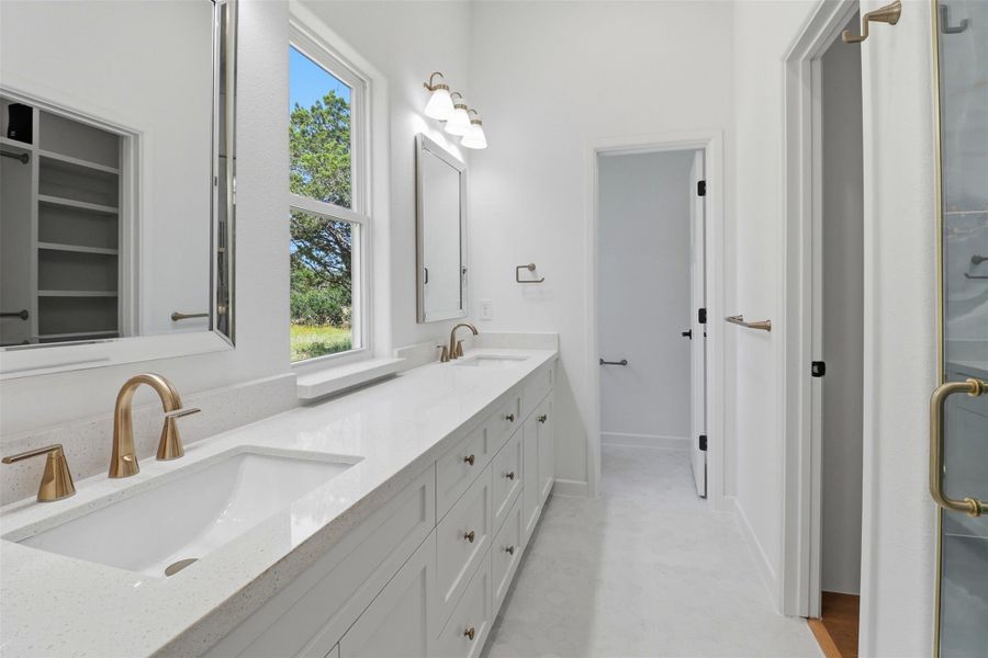 Dual vanity with a white countertop and integrated sinks, brushed brass hardware, framed mirrors, a window, and an adjacent shower enclosure Dual vanity with a white countertop and integrated sinks, brushed brass hardware, framed mirrors, a window, and an adjacent shower enclosure