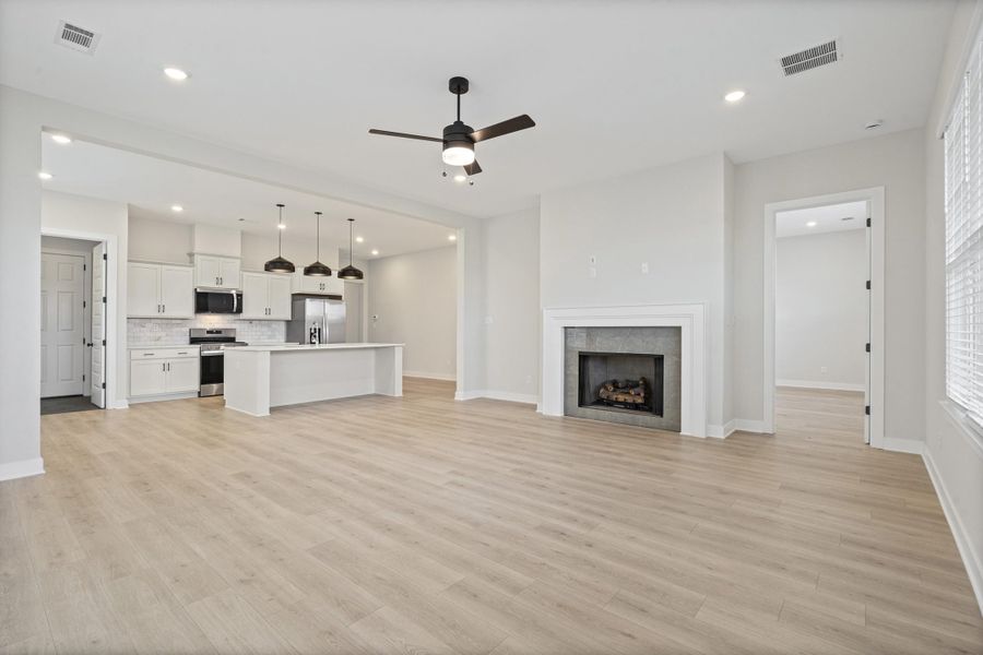 Open-concept living space featuring wood-finish flooring, a contemporary fireplace with a white mantel, and a kitchen island with pendant lighting Open-concept living space featuring wood-finish flooring, a contemporary fireplace with a white mantel, and a kitchen island with pendant lighting