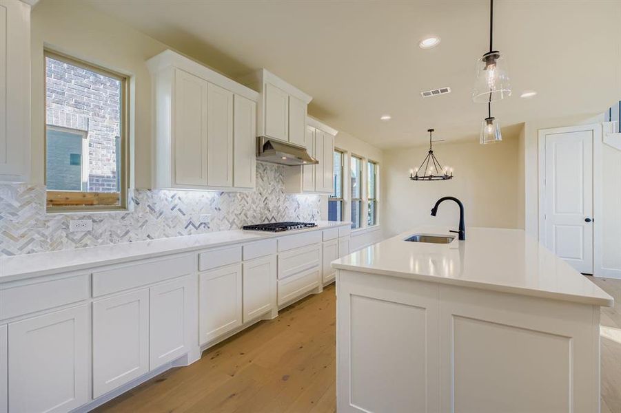 Kitchen featuring tasteful backsplash, white cabinetry, light wood finished floors, pendant lighting, and a kitchen island with sink