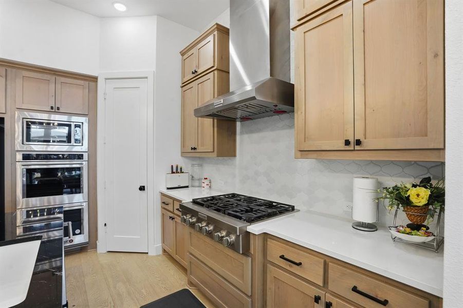 Kitchen featuring backsplash, stainless steel appliances, wall chimney exhaust hood, light wood-type flooring, and light stone counters Kitchen featuring backsplash, stainless steel appliances, wall chimney exhaust hood, light wood-type flooring, and light stone counters