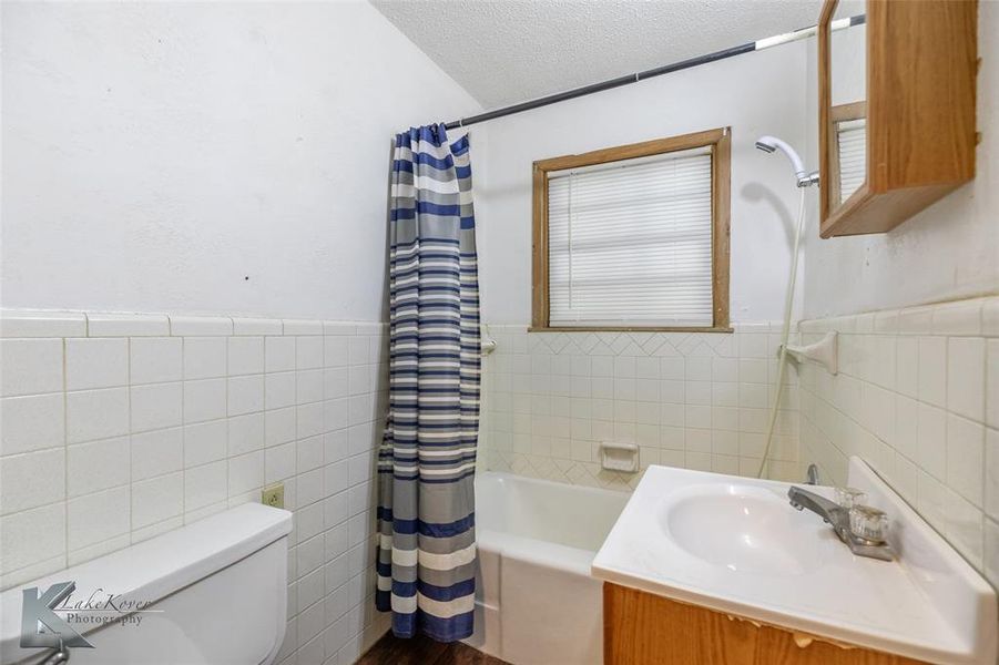 Full bathroom with a textured ceiling, tile walls, wainscoting, vanity, and shower / tub combo