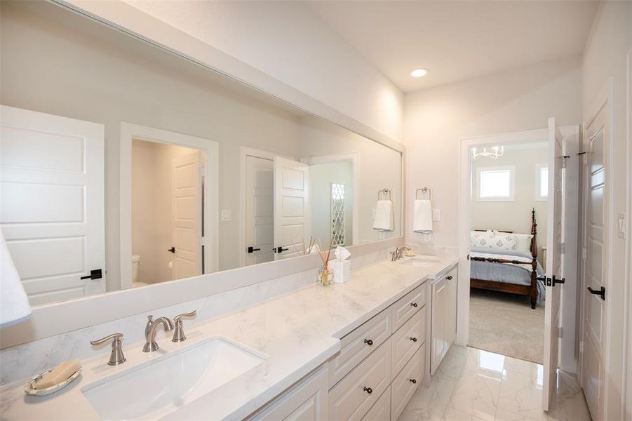 This photo showcases a modern bathroom with a long marble countertop featuring dual sinks and elegant fixtures. The space is bright, with ample lighting and a large mirror. Adjacent is a glimpse of a bedroom with plush carpeting, creating a seamless and inviting transition between the spaces.