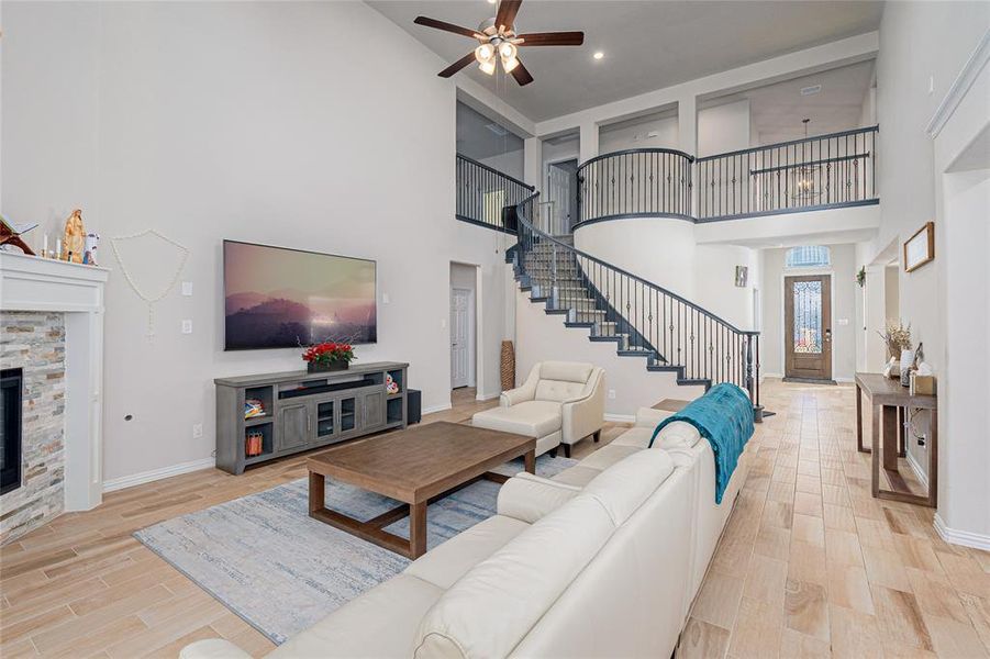 Living room with a stone fireplace, ceiling fan, a high ceiling, and light wood-type flooring