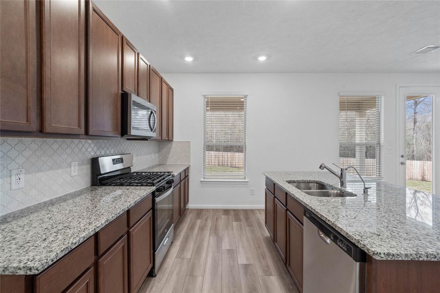 This spacious kitchen boasts soaring ceilings, Woodmont Cody cabinets in a Ginger stain, Granite Luna Pearl countertops, stainless steel Whirlpool appliances, a modern Daltile backsplash, recessed lighting, and a pantry—all overlooking the expansive Family Room.