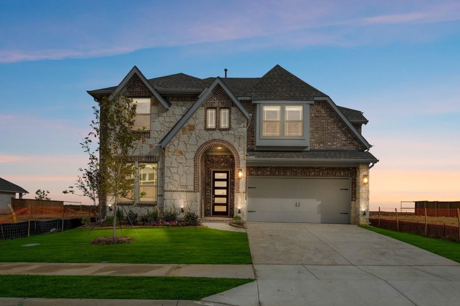 Two-story home with stone and brick exterior, arched entry, two-car garage, and green lawn at dusk